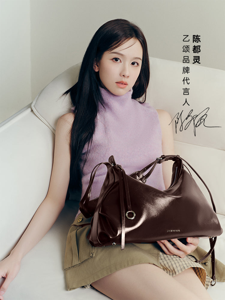 Woman in lavender sleeveless top posing with glossy dark brown Amazing Song handbag on sofa