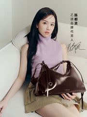 Woman in lavender sleeveless top posing with glossy dark brown Amazing Song handbag on sofa