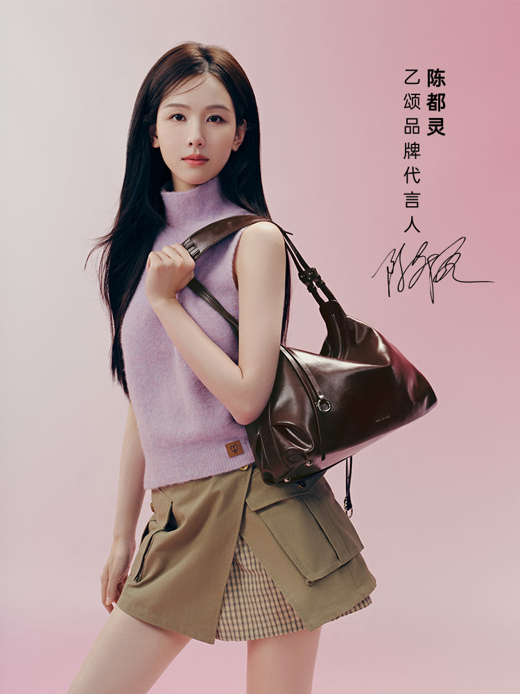 Woman in lilac sleeveless sweater and khaki skirt posing with brown shoulder bag on pink background