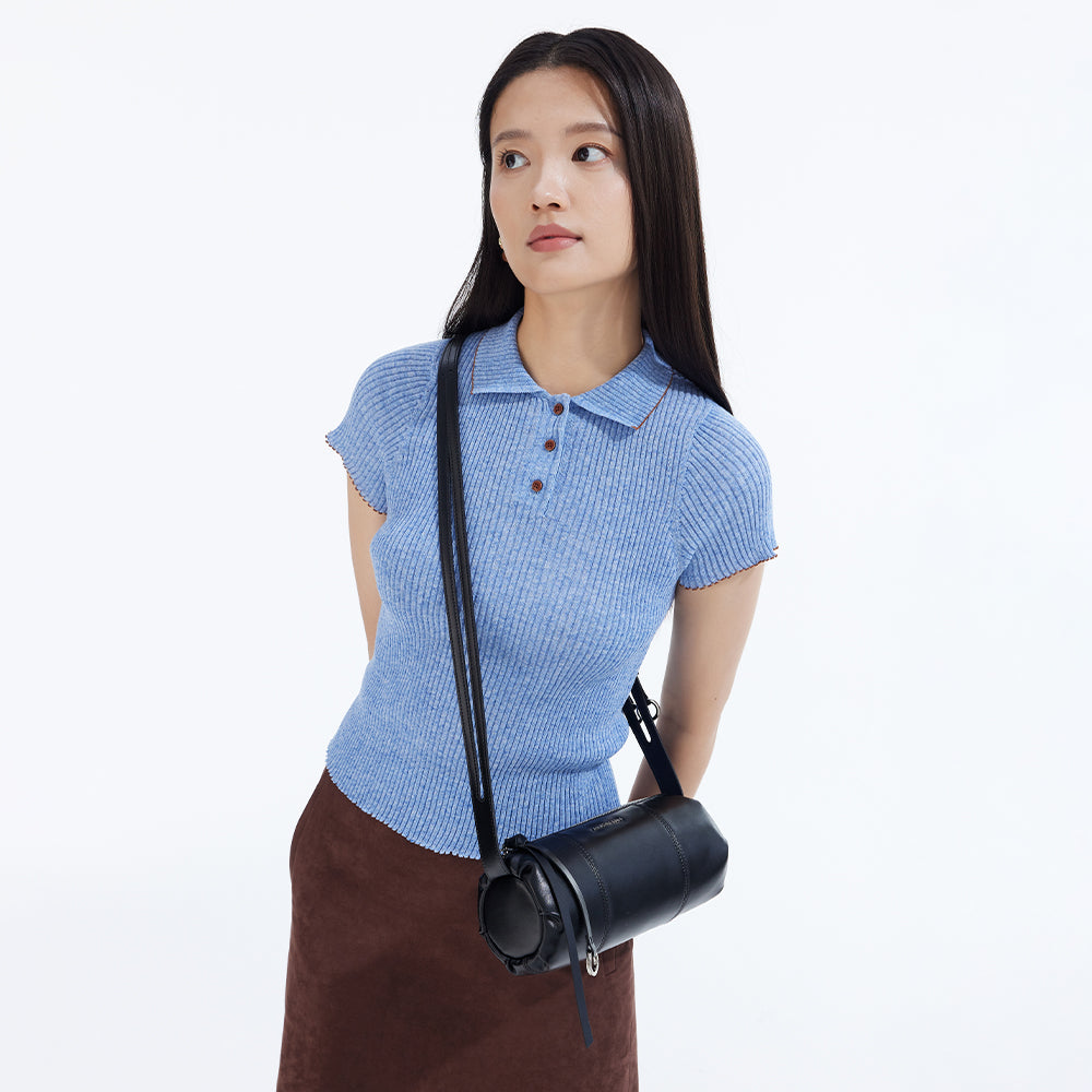 Woman in light blue ribbed polo top and brown skirt with black crossbody bag, white background