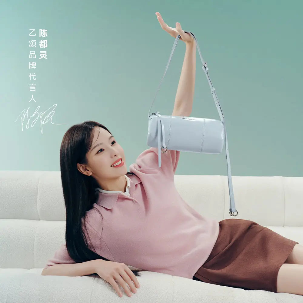 Smiling woman in pink top holding pastel blue handbag on white sofa, Amazing Song brand promo