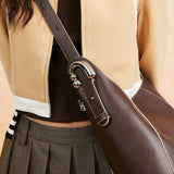Close-up of a woman wearing a brown leather shoulder bag with unique silver hardware, plaid skirt, and beige jacket, Amazing Song fashion style.