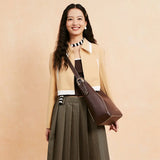 Smiling woman in beige jacket, plaid skirt, and brown crossbody bag against neutral background