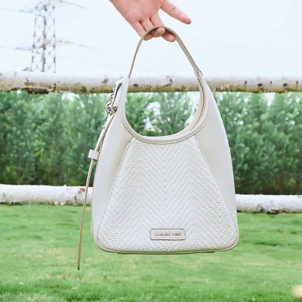 Hand holding woven cream Amazing Song handbag outdoors with greenery and fence