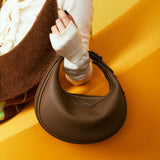 Brown crescent leather handbag held by woman in cozy sweater, Amazing Song official site