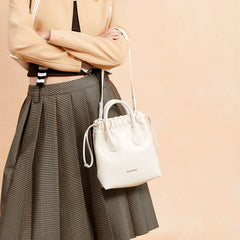 Woman in pleated skirt and beige jacket holding white Amazing Song bucket bag