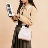 Woman playing melodica, wearing tan jacket, pink Amazing Song bag, and pleated skirt