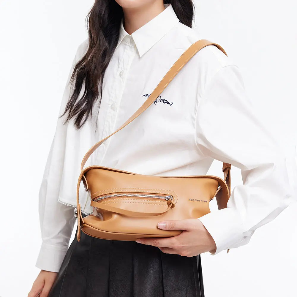 Woman in white shirt holding tan Amazing Song crossbody bag against white background