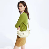 Woman in green sweater and tan skirt posing with cream crossbody bag on white background