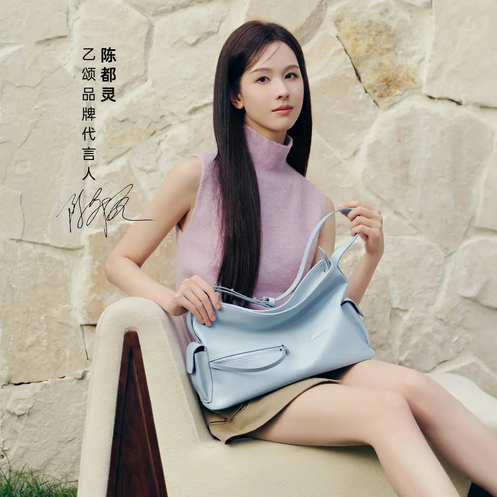 Woman in sleeveless lavender top holding a light blue handbag, sitting by a stone wall, Amazing Song