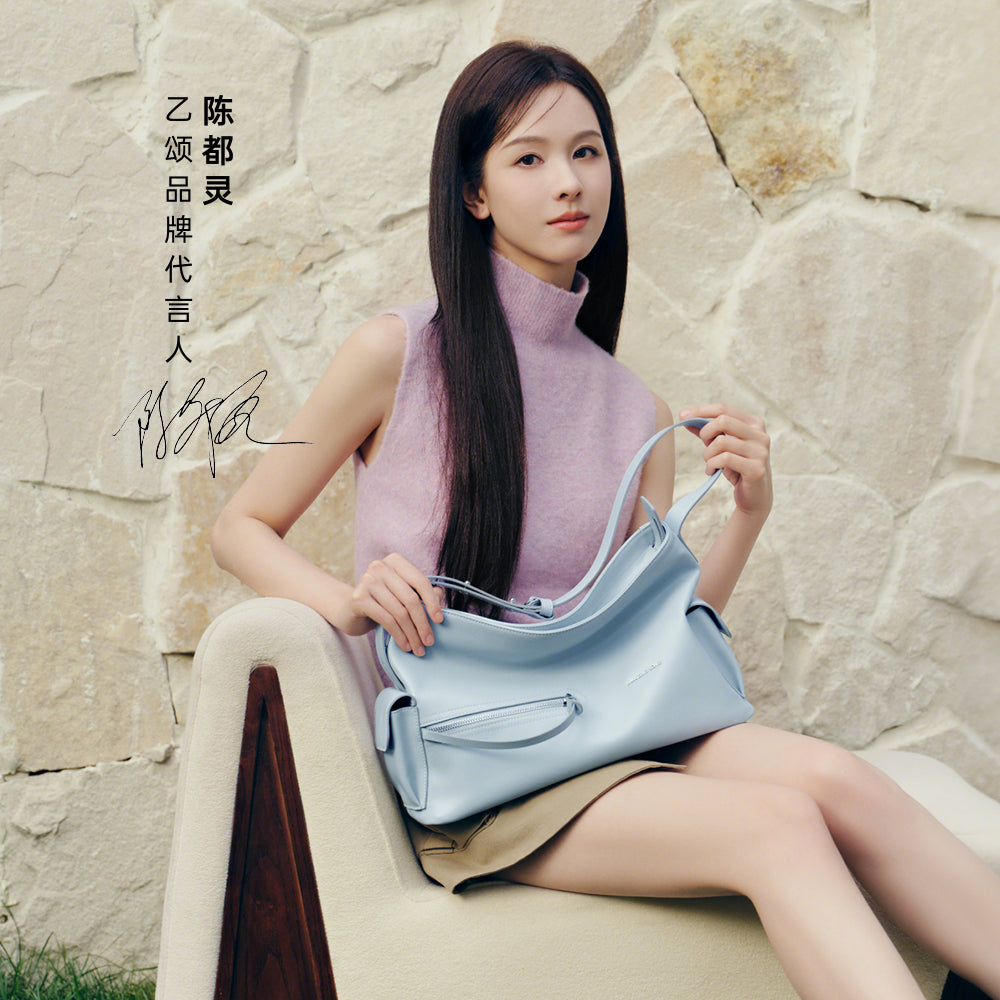 Woman in sleeveless lavender top holding a light blue handbag, sitting by a stone wall, Amazing Song