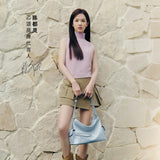Woman in sleeveless pink top and khaki skirt holding a light blue handbag, stone wall background