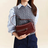 Woman holding a burgundy leather handbag, wearing a gray cable knit vest and blue shirt