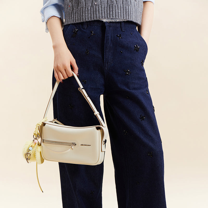 Woman holding cream Amazing Song handbag, wearing navy embroidered pants and grey sweater
