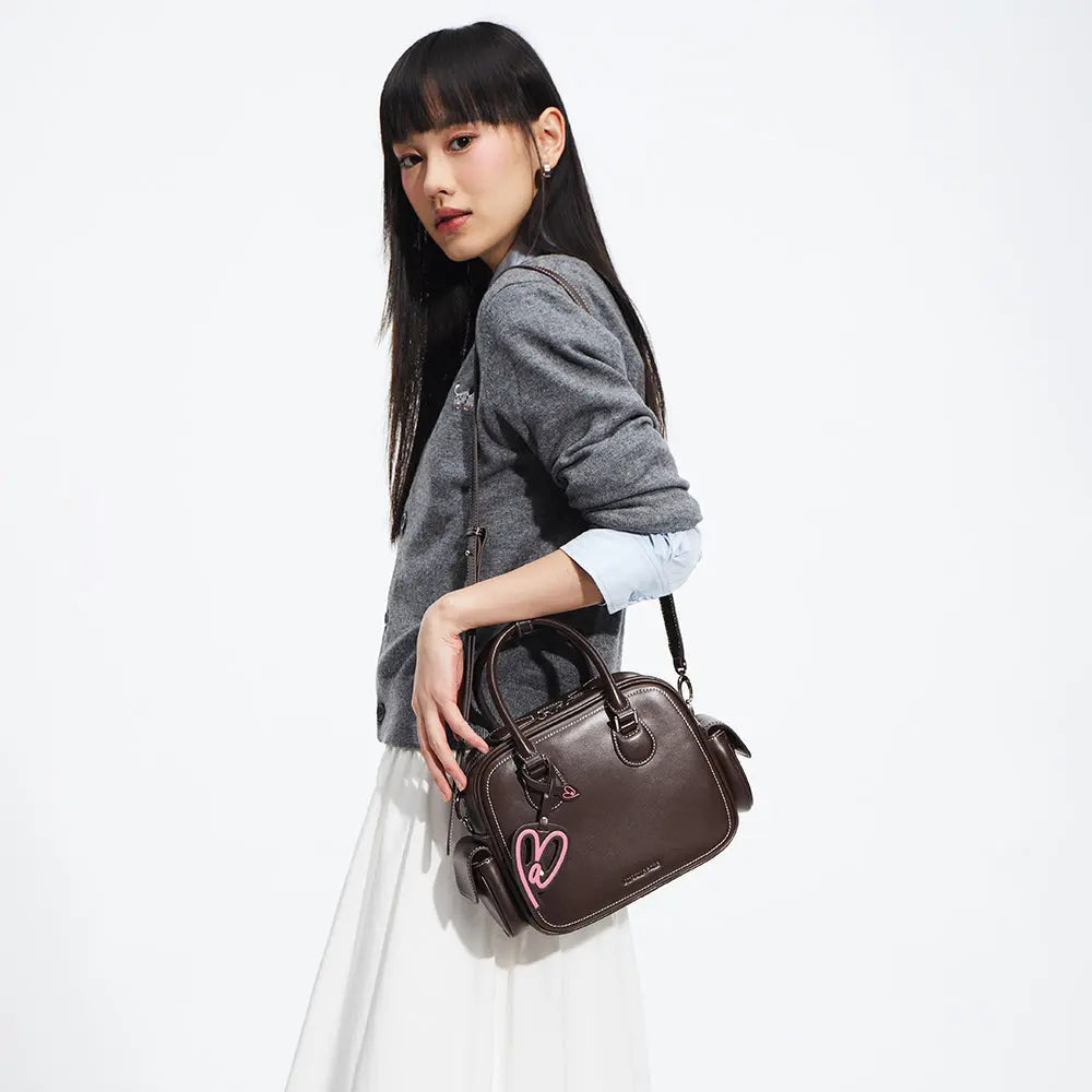 Woman in grey sweater holding dark brown handbag with pink heart charm, Amazing Song style