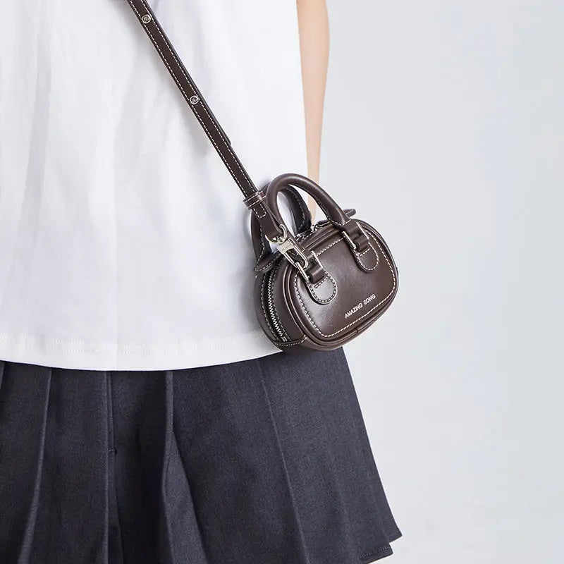 Dark brown mini crossbody handbag with 'Amazing Song' logo, worn over a white shirt and skirt.