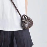 Dark brown mini crossbody handbag with 'Amazing Song' logo, worn over a white shirt and skirt.