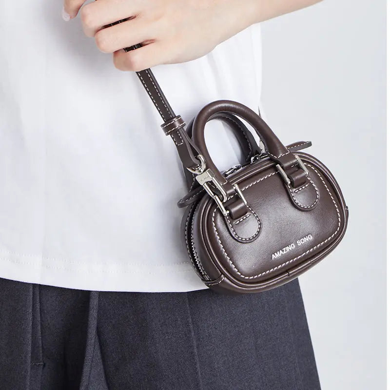 Hand holding small brown leather handbag with Amazing Song logo, white shirt background