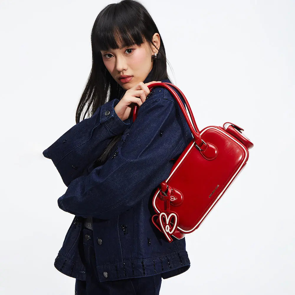 Woman in dark denim jacket posing with a red Amazing Song shoulder bag and heart keychain