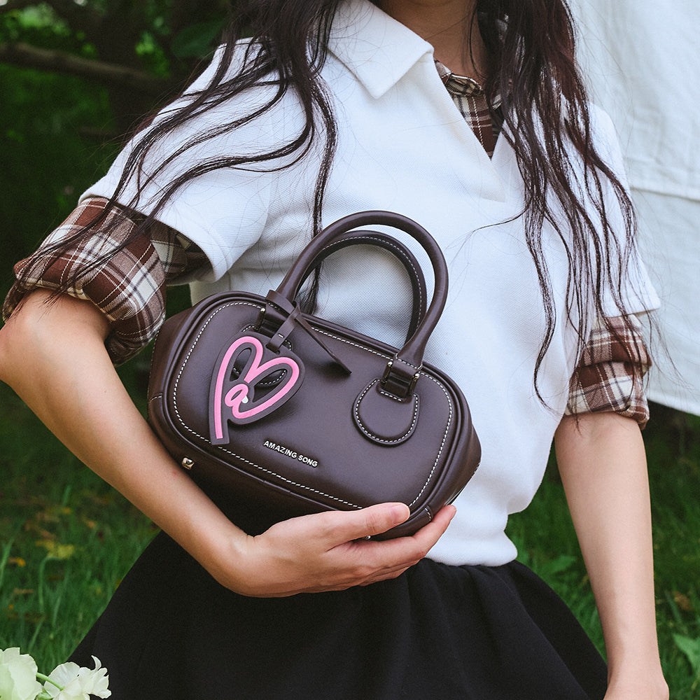 Woman holding brown Amazing Song handbag with pink heart charm outdoors