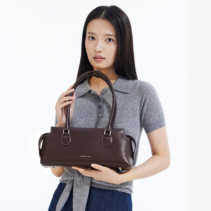 Woman holding brown leather Amazing Song handbag, grey polo sweater, minimal backdrop