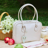 White leather handbag with charm on picnic blanket, apples, basket of flowers, outdoor setting