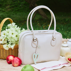 White leather handbag with charm on picnic blanket, apples, basket of flowers, outdoor setting