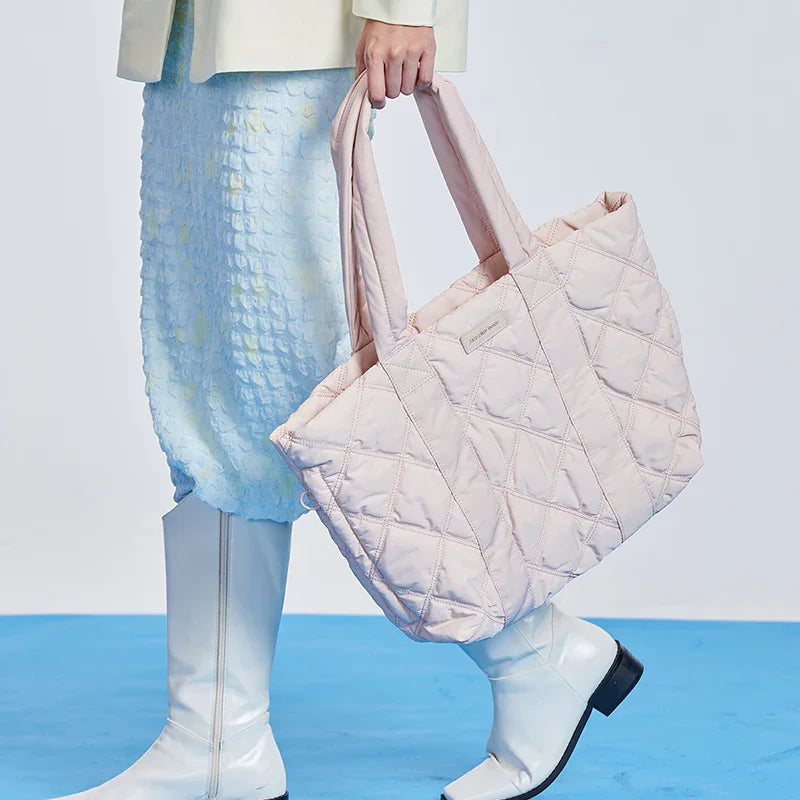 Woman holding a large quilted pink tote bag, wearing a pastel skirt and white boots