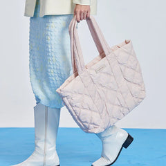 Woman holding a large quilted pink tote bag, wearing a pastel skirt and white boots