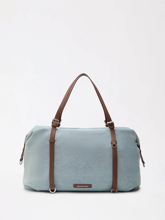 A donut-brown top-grain leather boston bag with rounded top and two short handles resting on a hotel bed