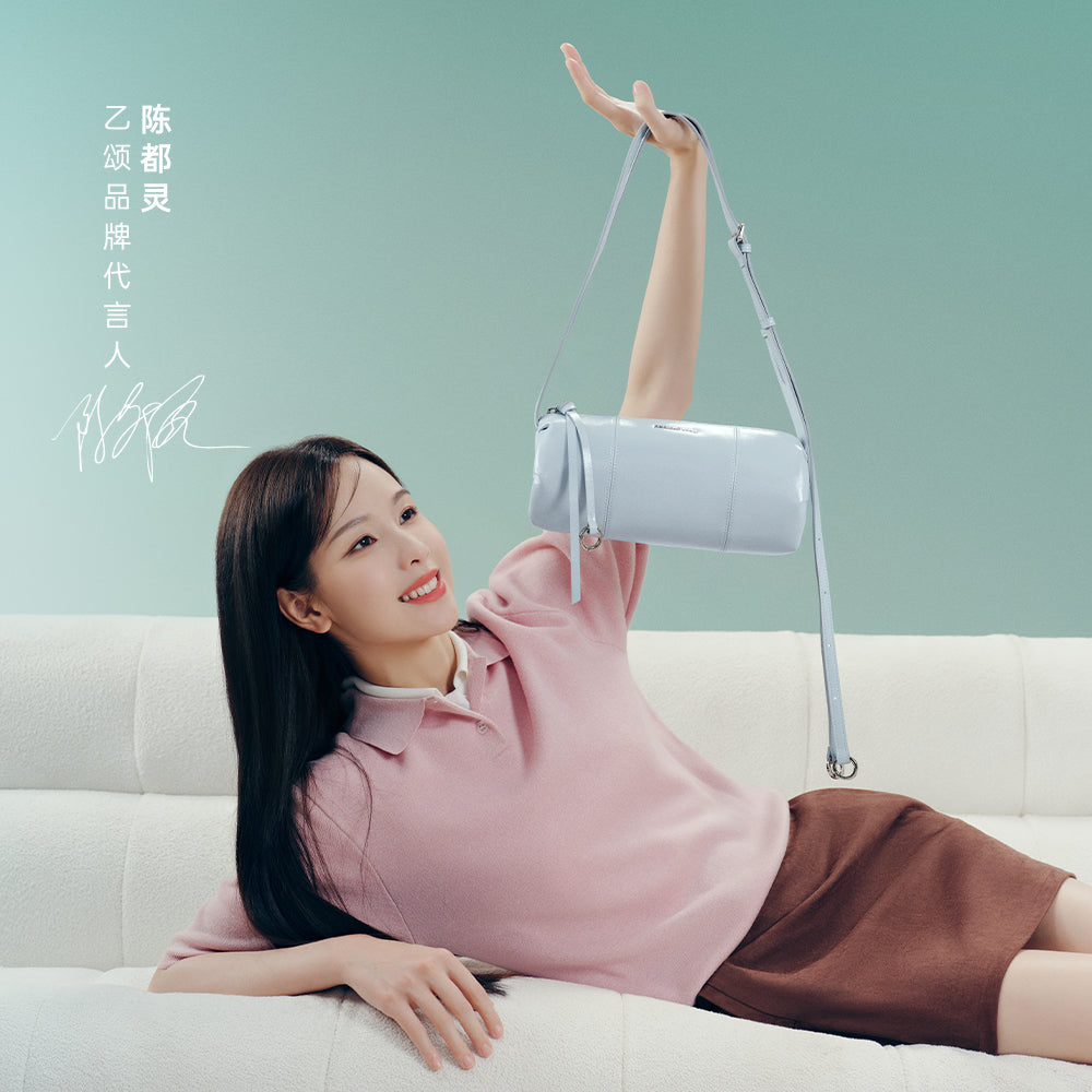 Smiling woman in pink top holding pastel blue handbag on white sofa, Amazing Song brand promo