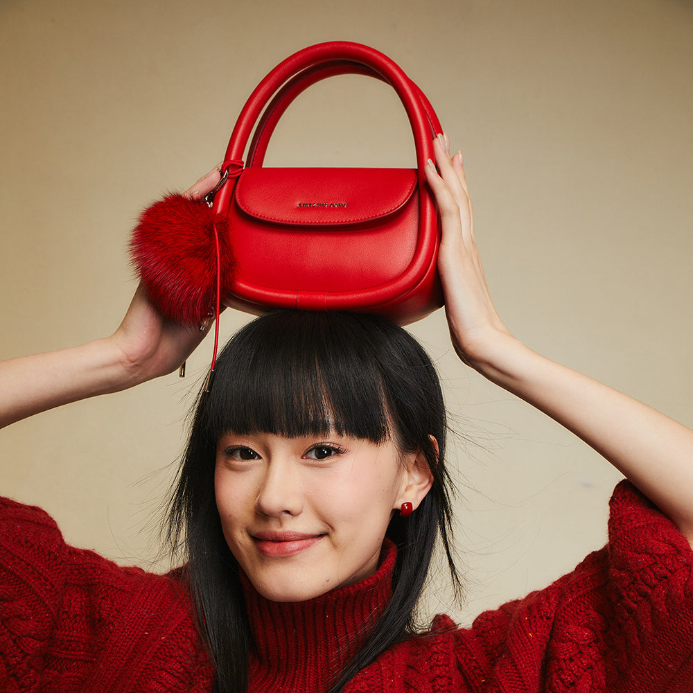Smiling woman in red sweater holding red handbag with pompom on head, Amazing Song brand
