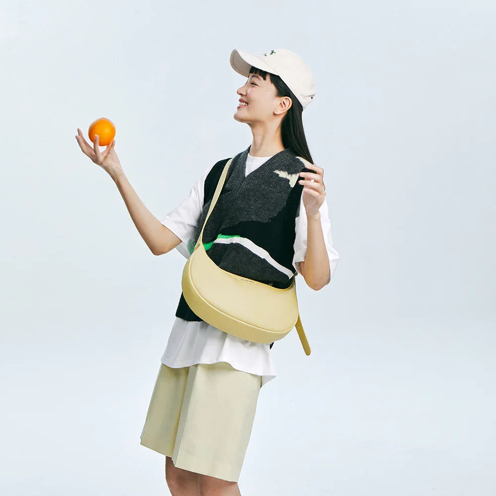 Smiling woman in a casual outfit with a light yellow sling bag holding an orange, Amazing Song official website