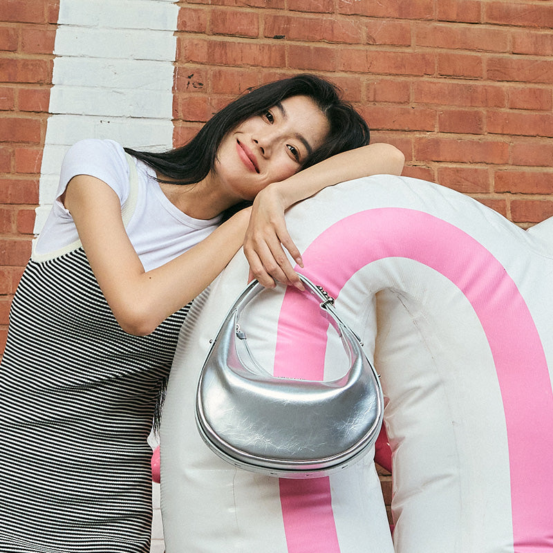 Smiling woman in striped dress holds silver handbag, leaning on pink and white inflatable, brick wall background.