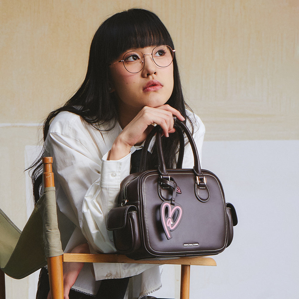 Woman with glasses in white shirt holding brown Amazing Song handbag with pink heart charm