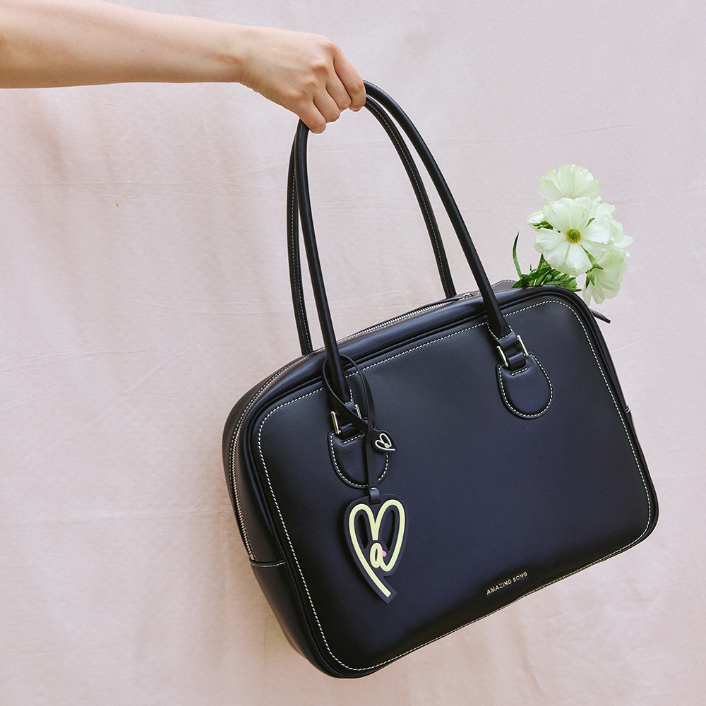 Hand holding black Amazing Song handbag with heart tag and white flowers against light background